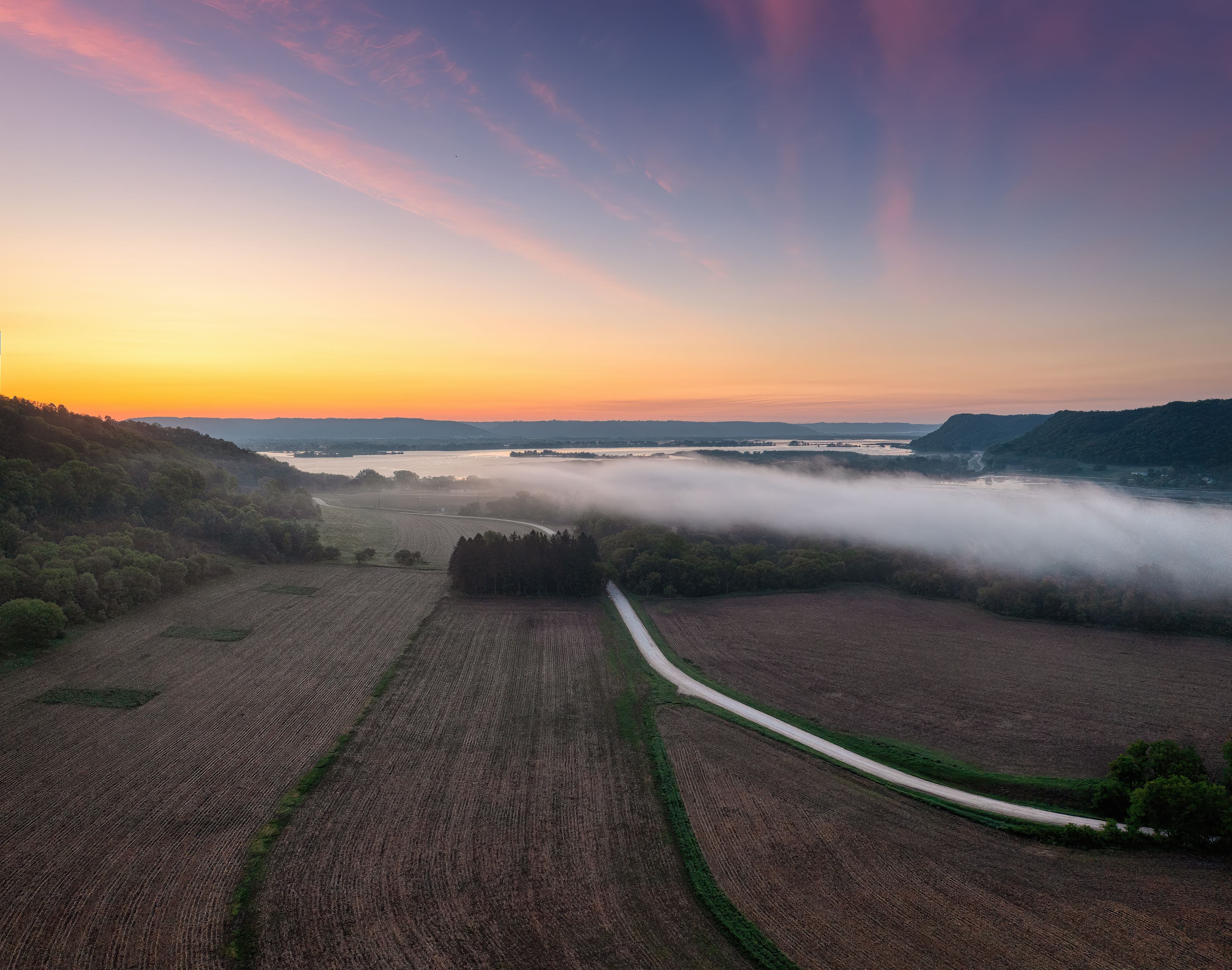 Aerial photograph of open country fields and a winding rural road at sunset.